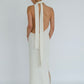 A woman viewed from the back wearing a white textured halter-neck midi dress with a long tie-back ribbon and a side slit, paired with black pointed heels against a light gray studio background.