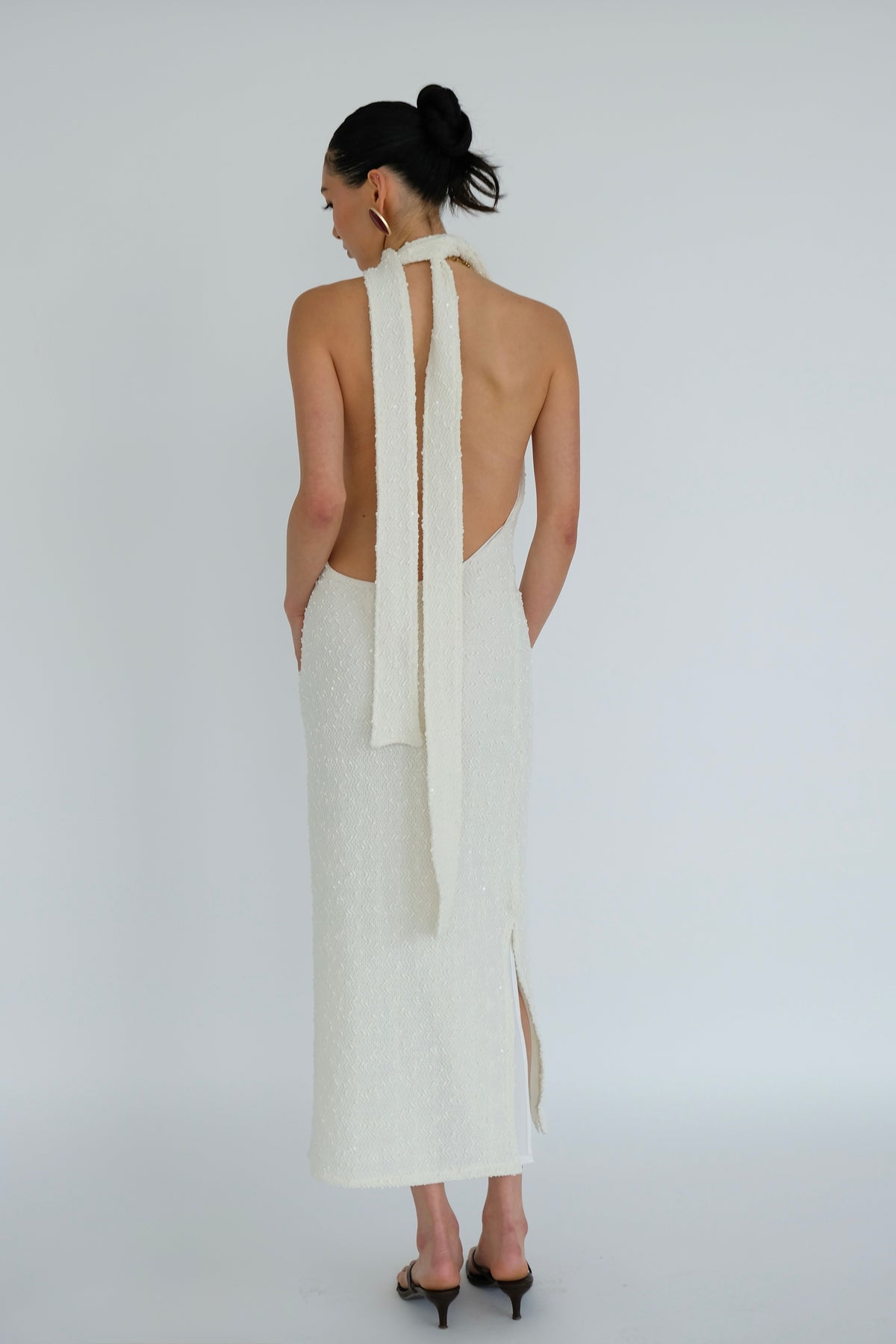 A woman viewed from the back wearing a white textured halter-neck midi dress with a long tie-back ribbon and a side slit, paired with black pointed heels against a light gray studio background.