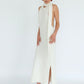 A model wearing a textured off-white halter-neck midi dress with side cutouts and a high slit, paired with black heeled sandals, set against a plain white studio background.