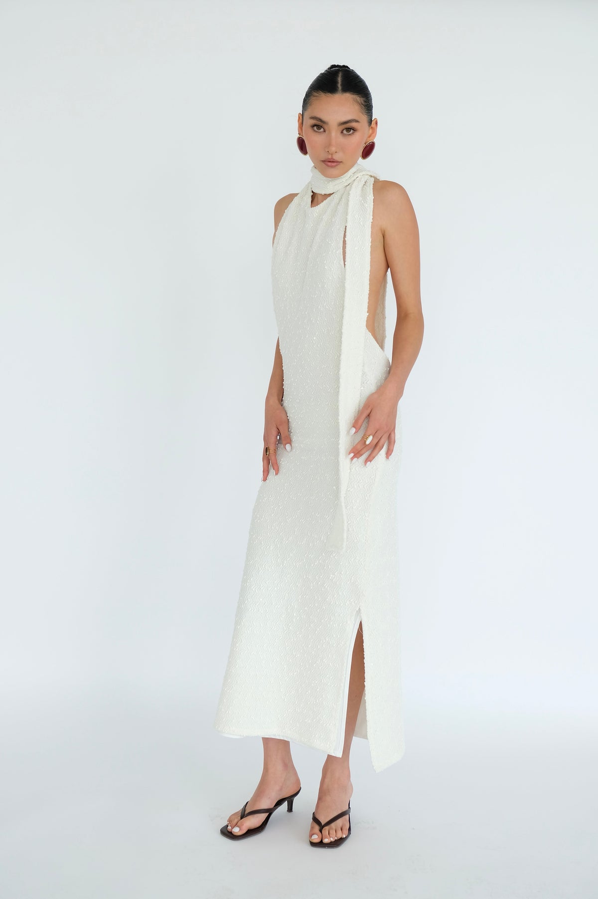 A model wearing a textured off-white halter-neck midi dress with side cutouts and a high slit, paired with black heeled sandals, set against a plain white studio background.