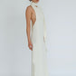 A model wearing a long, sleeveless white dress with a high draped collar and side slit, paired with black heeled sandals, photographed against a seamless light gray studio background.