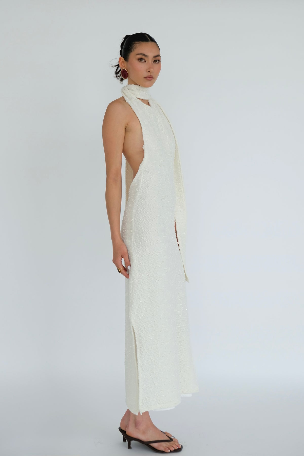 A model wearing a long, sleeveless white dress with a high draped collar and side slit, paired with black heeled sandals, photographed against a seamless light gray studio background.